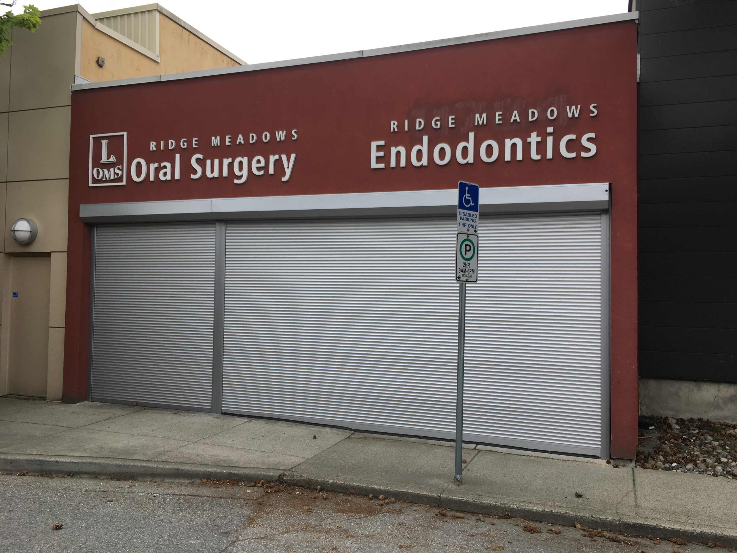 A storefront with signs for Ridge Meadows Oral Surgery and Ridge Meadows Endodontics. The storefront has two large metal roll-up doors that are closed. There is a sign in front indicating a disabled parking spot.