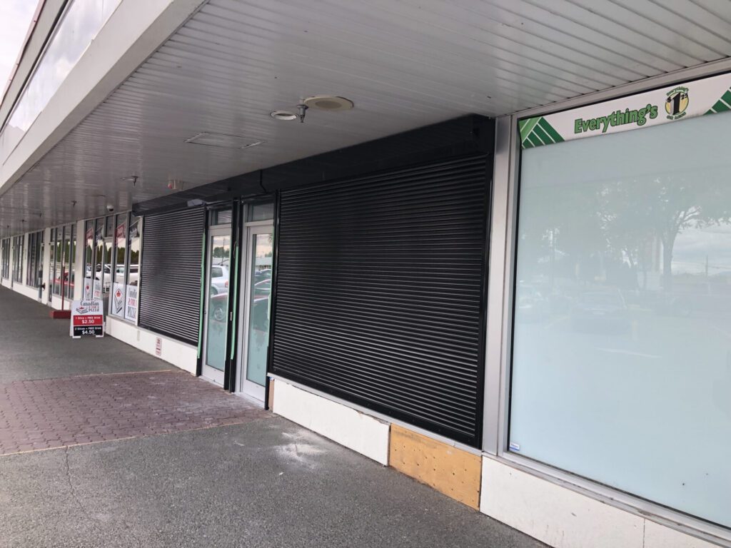 A row of storefronts with closed black shutters and a sign for "Everything's" on the right. A small sign for "Canadian 2 for 1 Pizza" is visible in front of one of the stores.