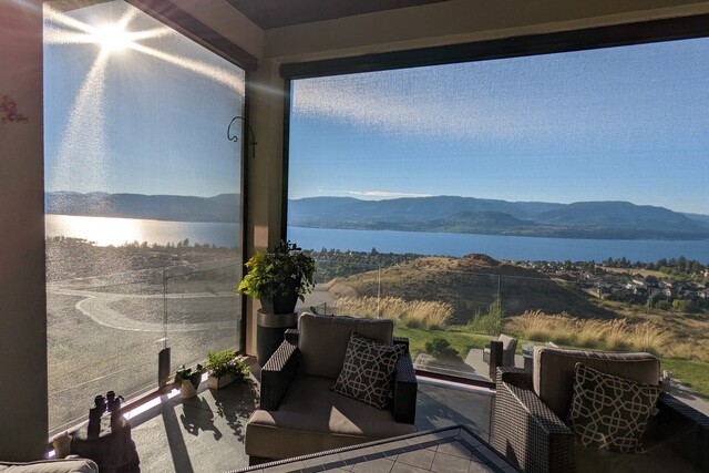 An outdoor patio with comfortable seating, overlooking a scenic view of a lake and distant mountains under a clear blue sky. A sun shade screen partially covers the patio, allowing light to filter through while providing shade and a cozy, sheltered atmosphere